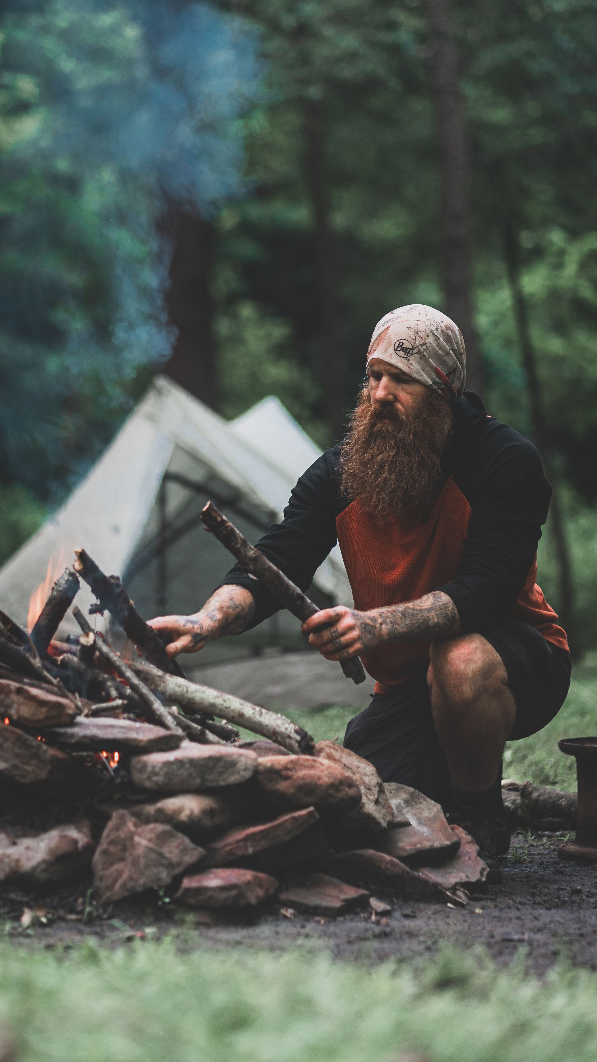 A man with a beard is kneeling next to a campfire in the woods.
