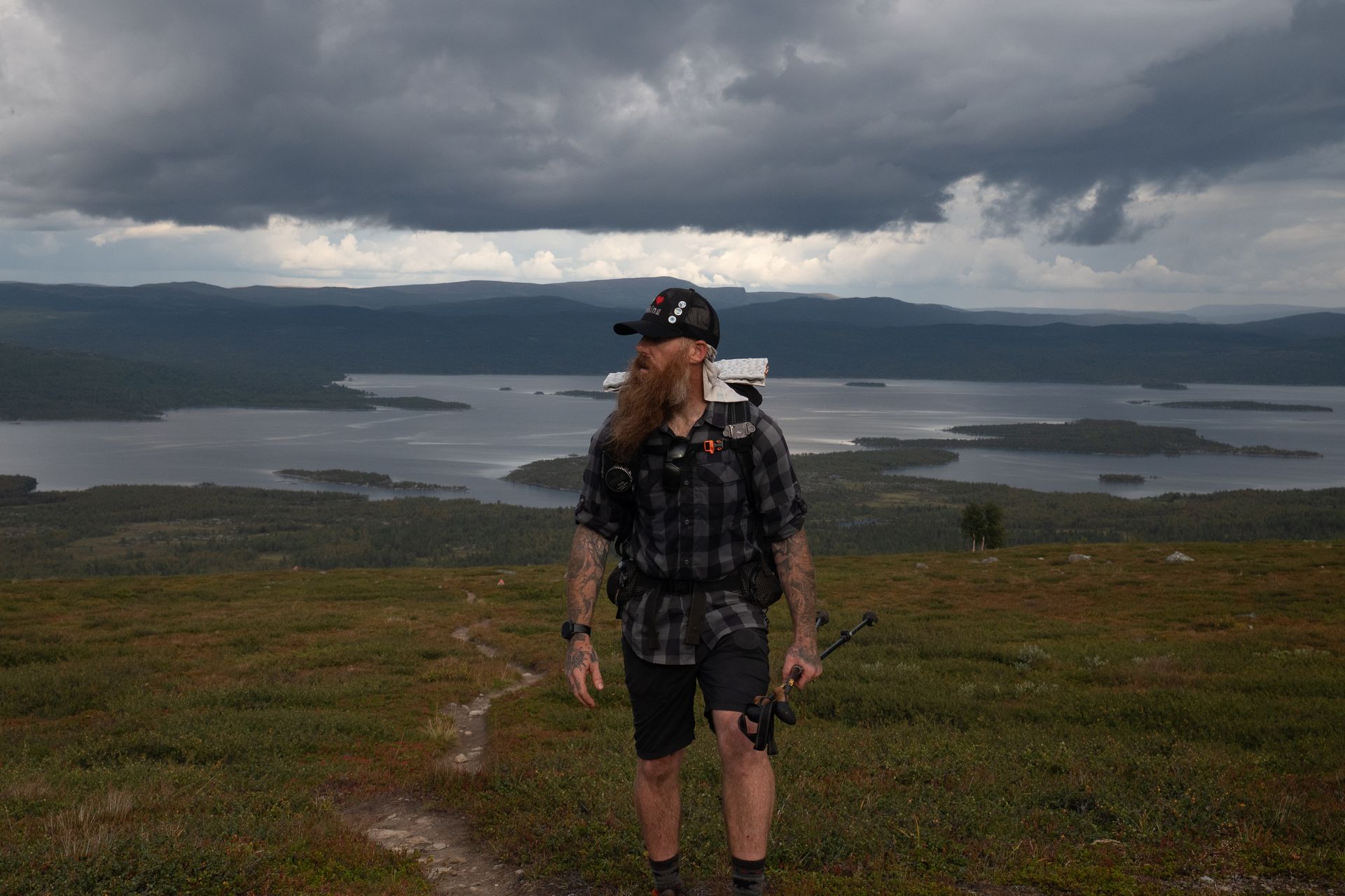 A man with a beard is standing on top of a hill overlooking a lake.