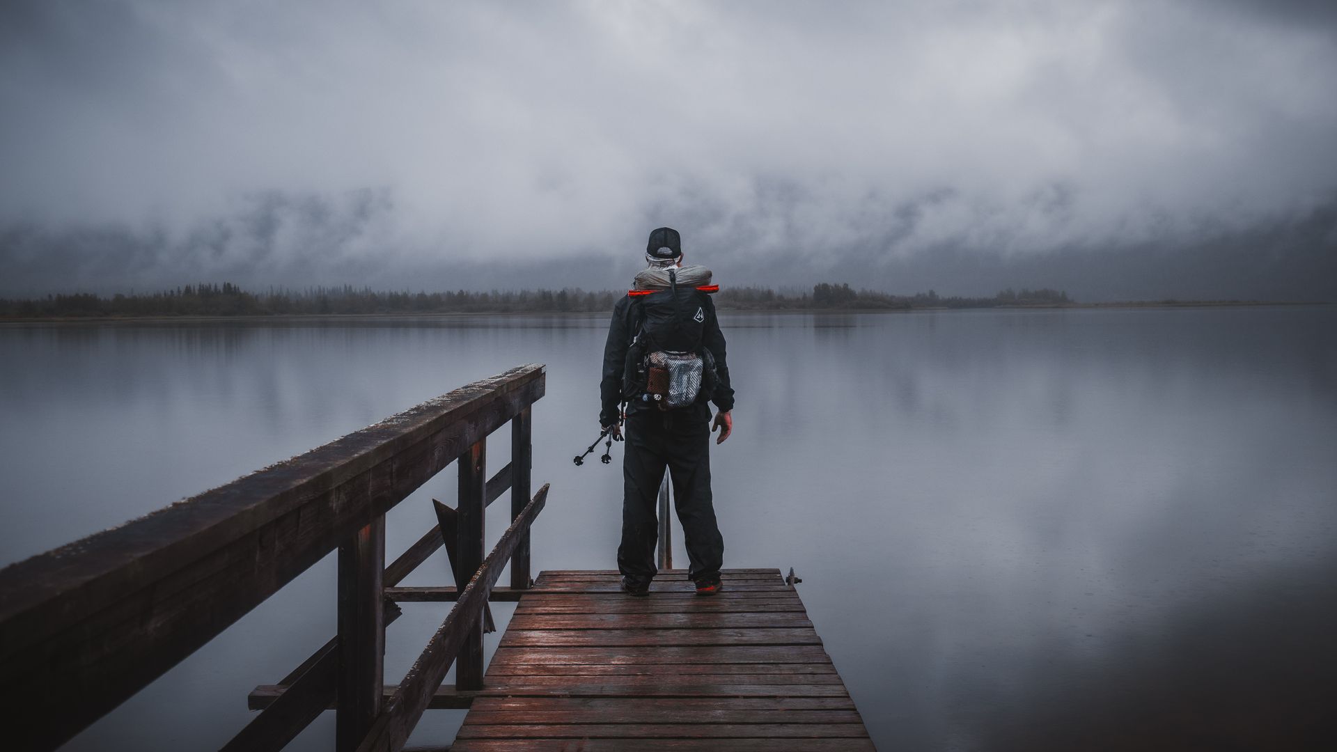 A man with a backpack is standing on a dock overlooking a lake.