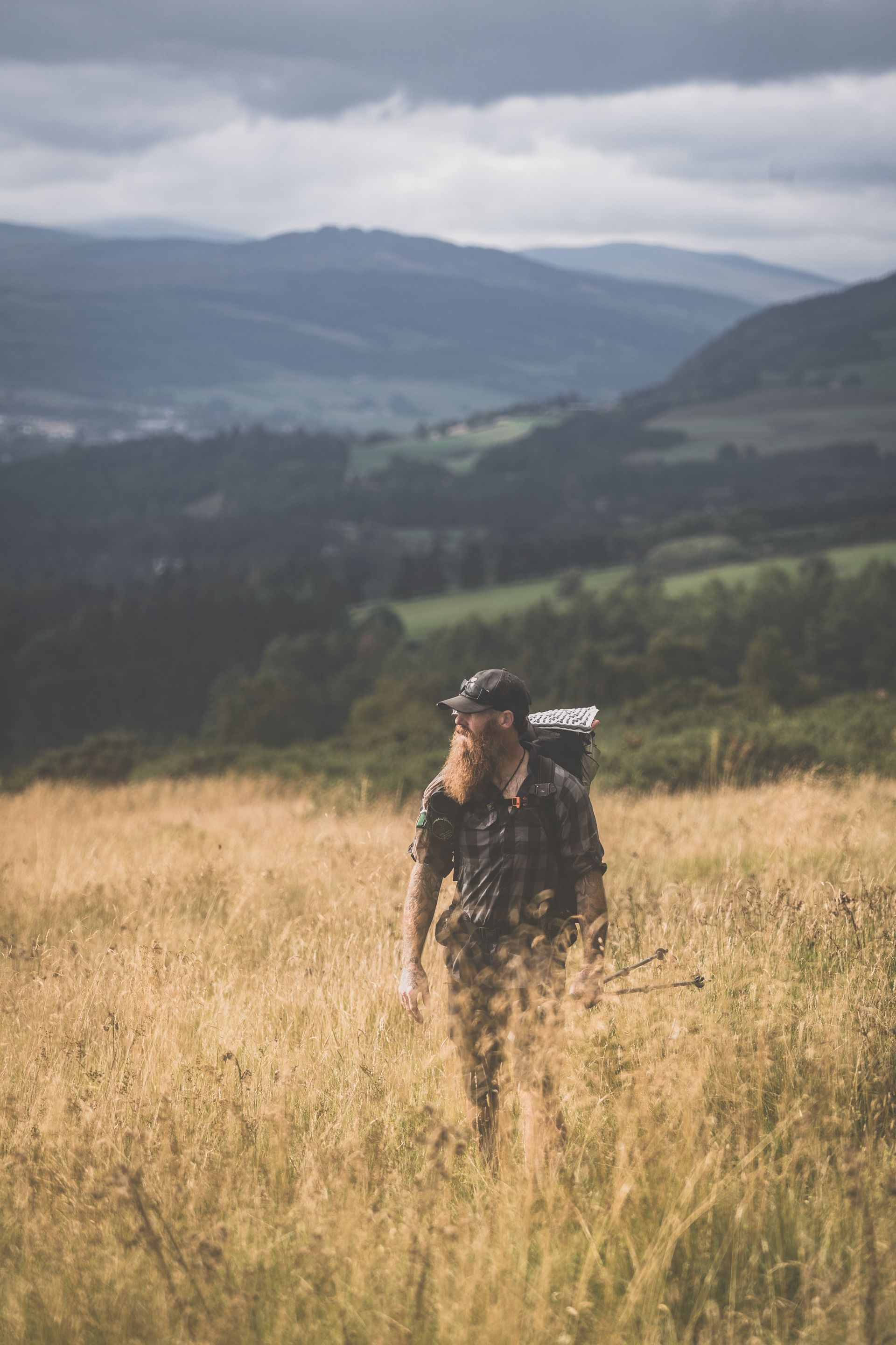 A man with a backpack is walking through a field with mountains in the background.