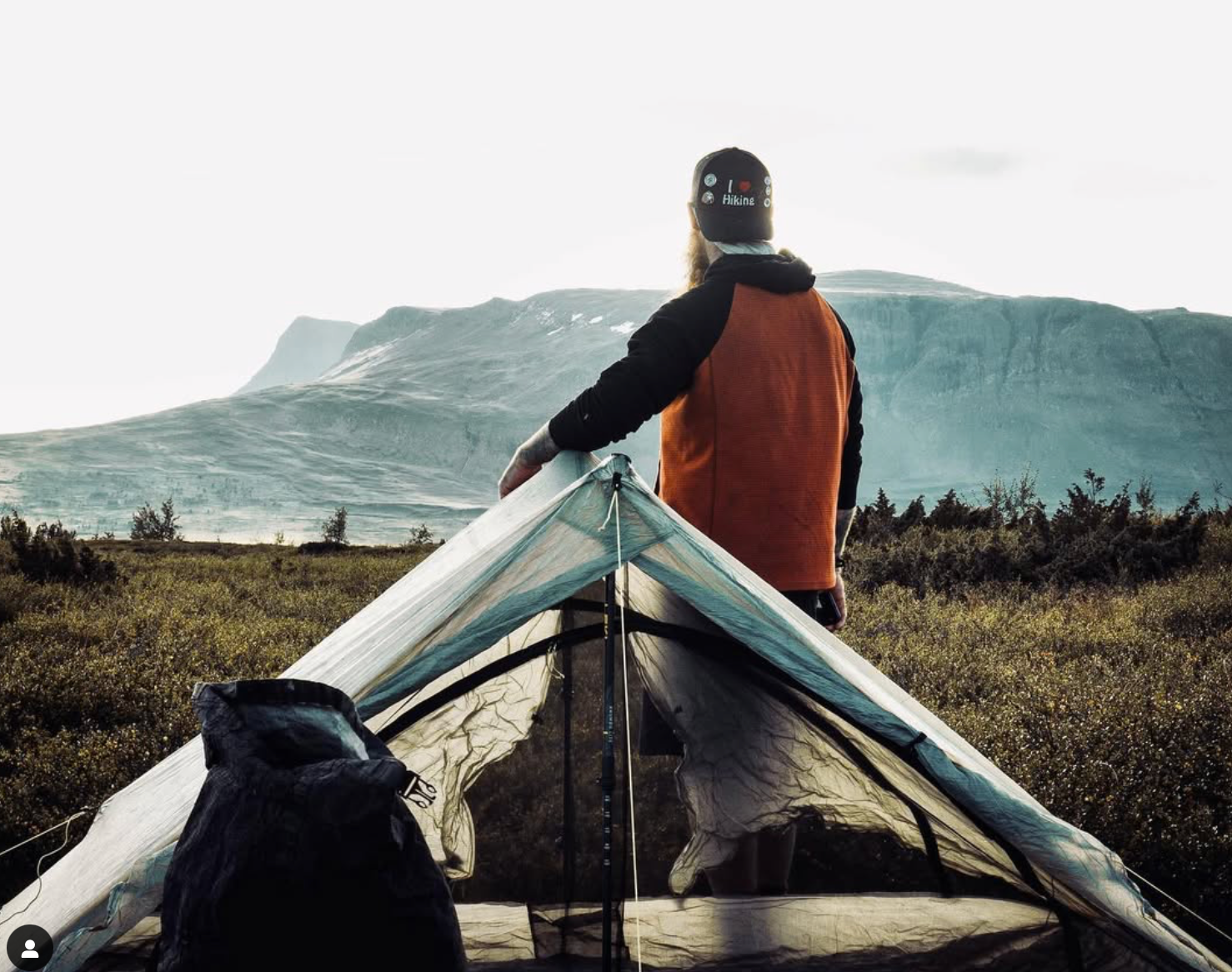 A man is standing in front of a tent with mountains in the background