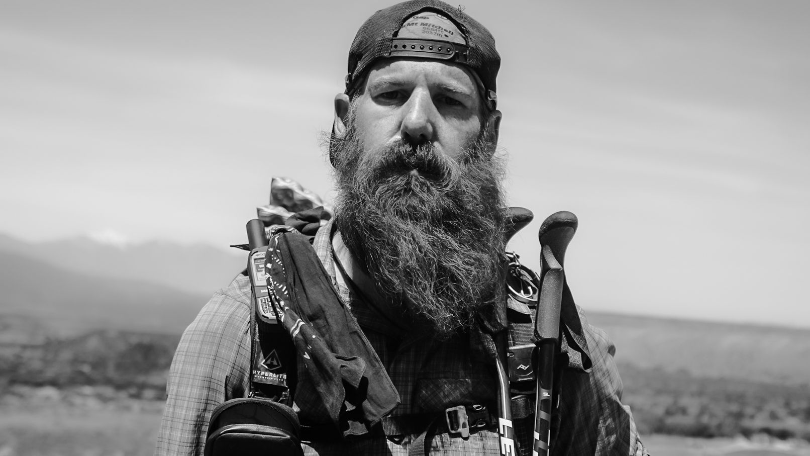 A black and white photo of a man with a beard holding hiking poles.