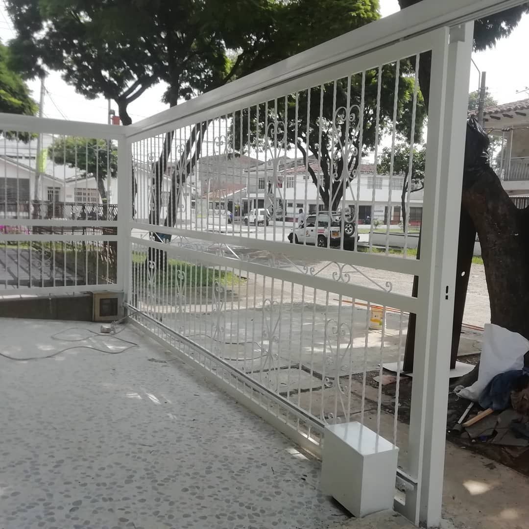 Puerta de metal blanco con barrotes verticales junto a una acera y un árbol en un entorno urbano.
