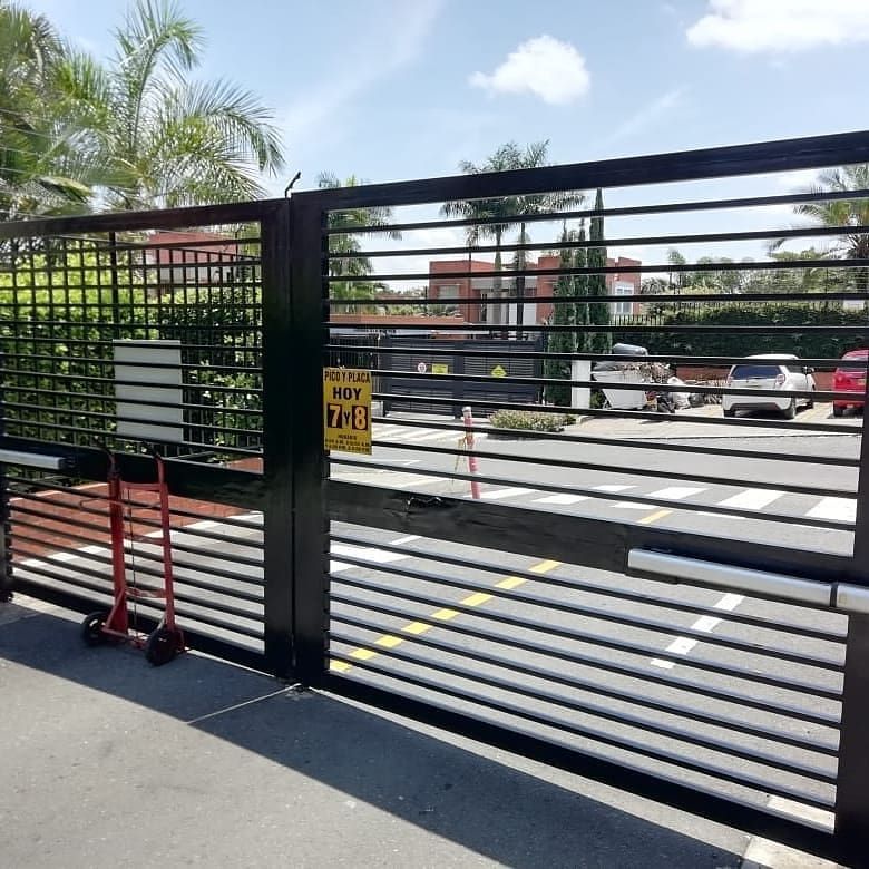 Una verja metálica negra con barrotes horizontales da acceso desde la calle, con un cartel amarillo y coches aparcados más allá.
