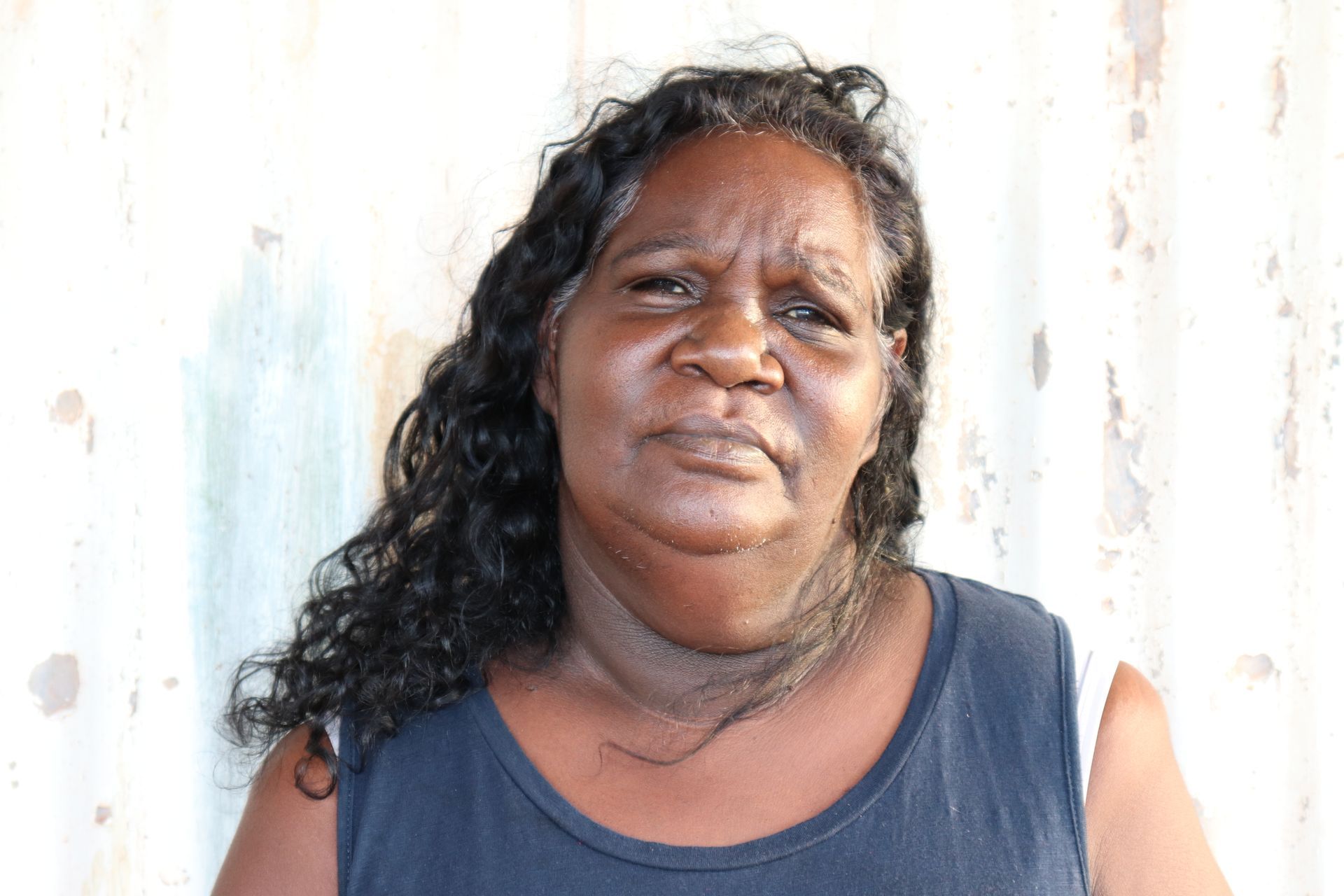A woman with long hair and a black tank top is standing in front of a white wall.