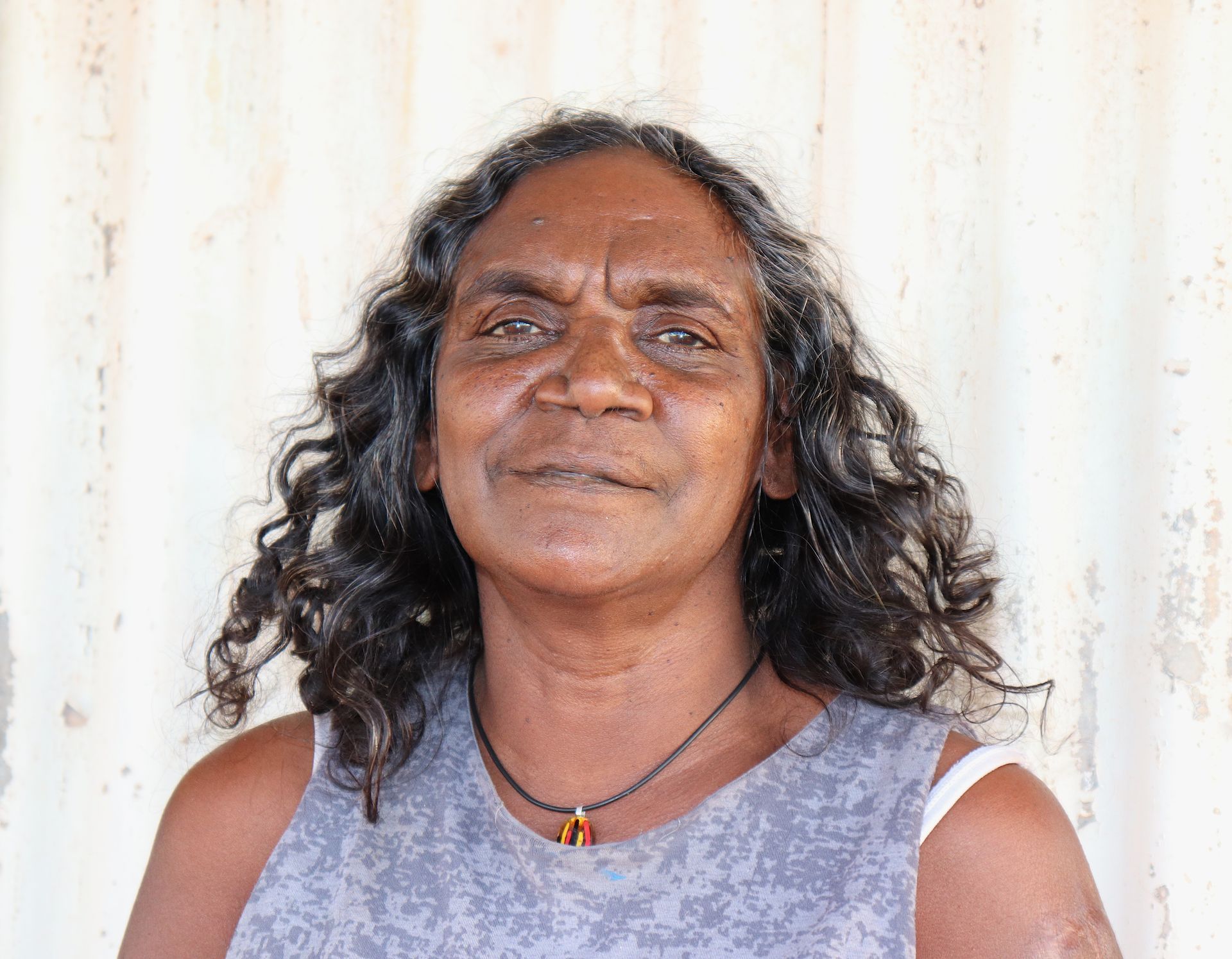 A woman with long curly hair and a necklace is standing in front of a wall.