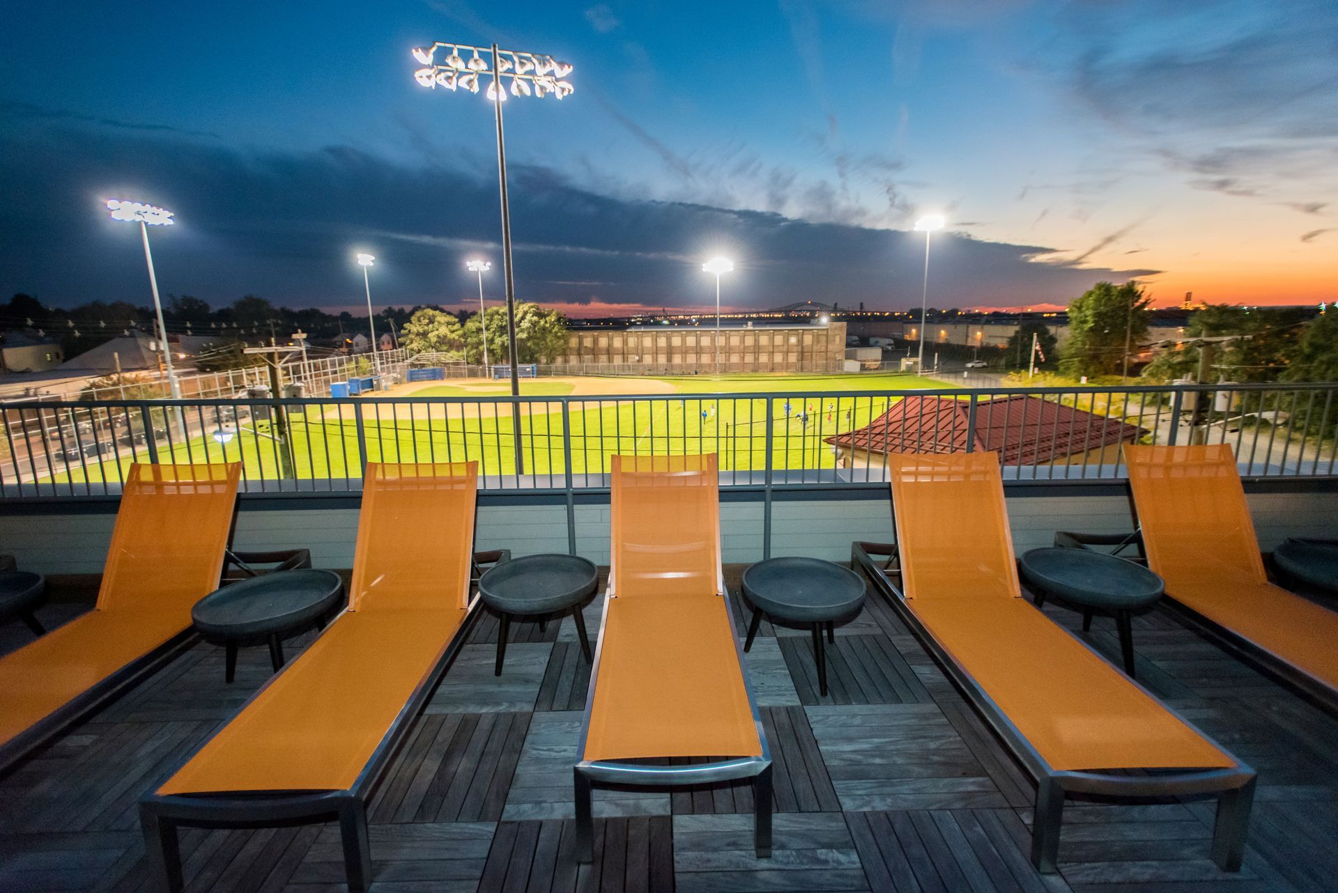 A row of lounge chairs on a rooftop with a view of a baseball field.