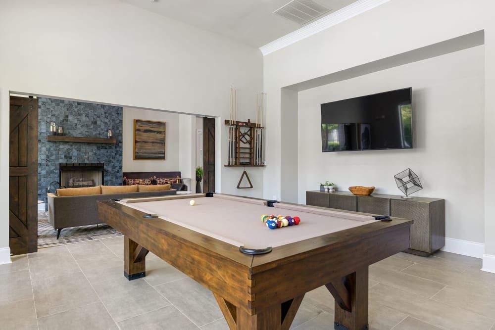 Modern game room featuring a pool table with racked balls in the foreground, a seating area with a stylish fireplace, and a wall-mounted flat-screen TV. The room has neutral walls, tiled flooring, and is well-lit by natural light at Marquis Carmel Commons in Charlotte, NC.