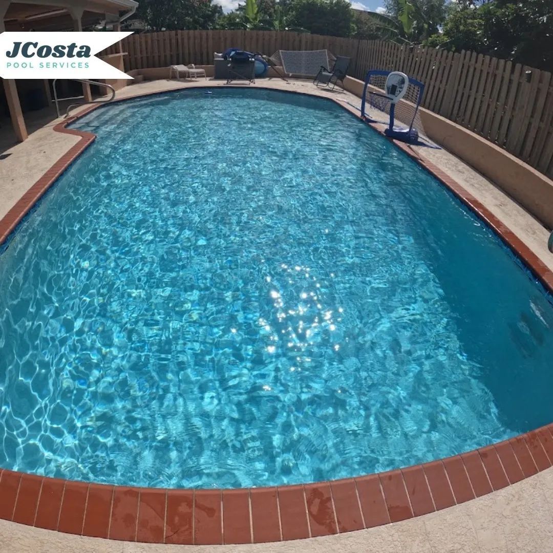 A turquoise pool in a backyard, with a brick border and a fence in the background.