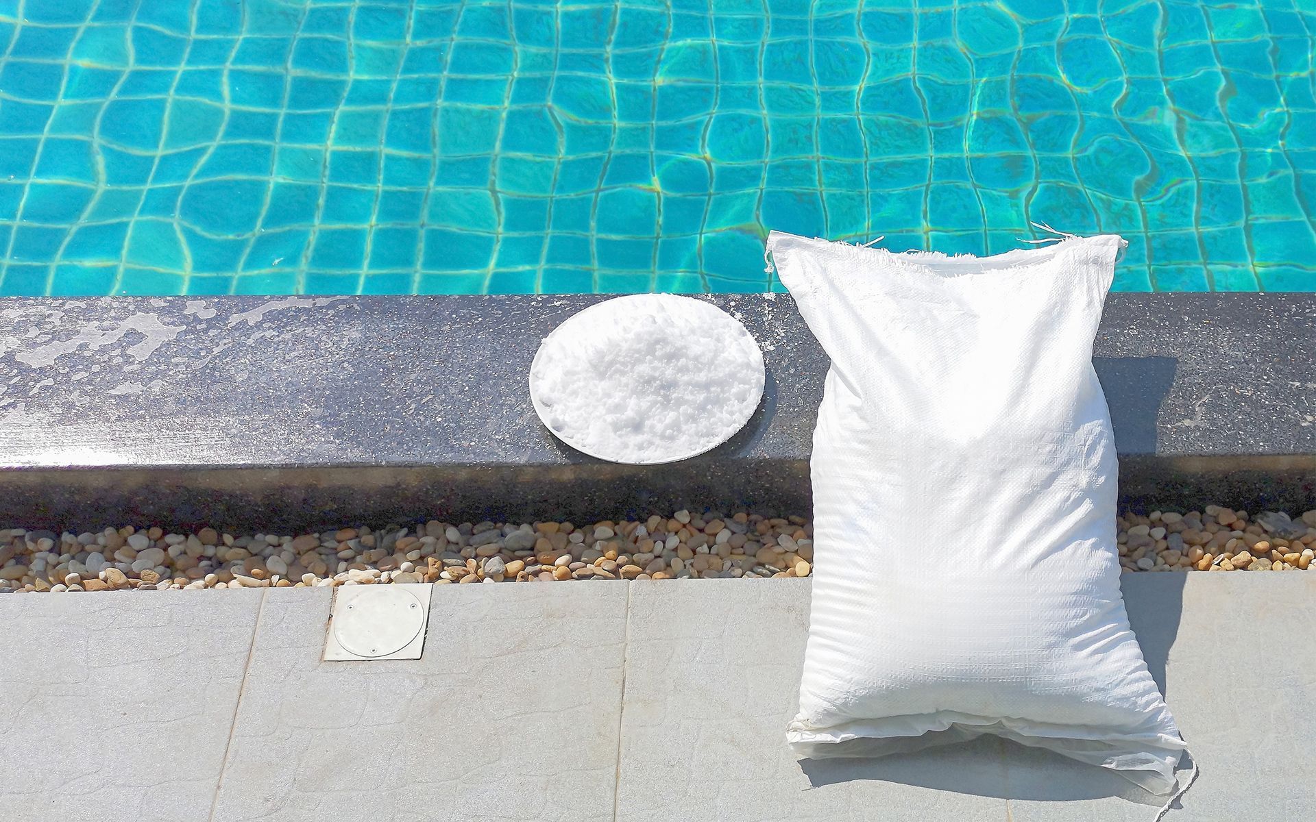 Bag and pile of white pool salt next to a turquoise swimming pool.