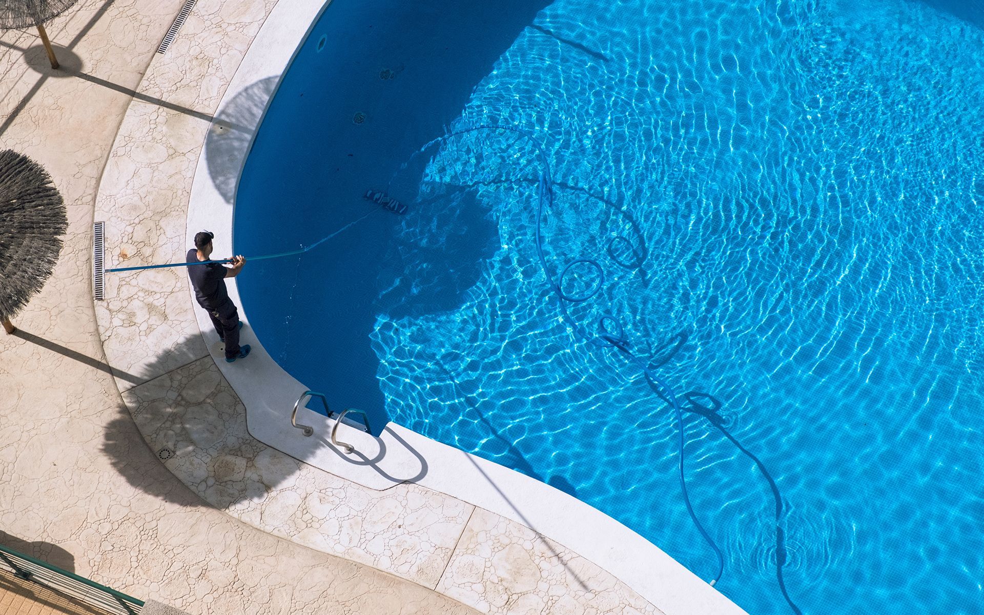 Man cleaning a bright blue swimming pool with a long-handled brush, outdoors on a sunny day.