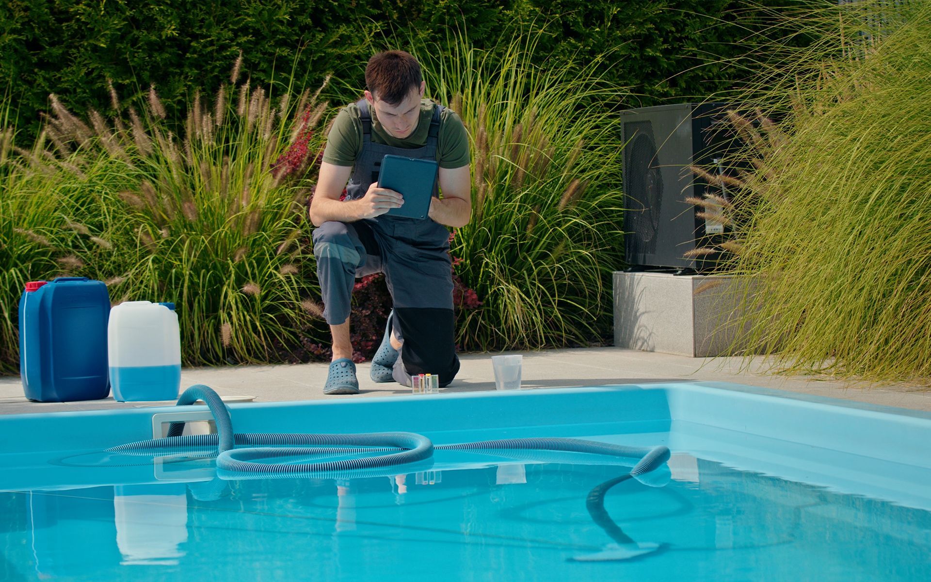 Man kneeling at pool's edge, testing water. Tall grass and chemicals are nearby. Blue water, sunny day.