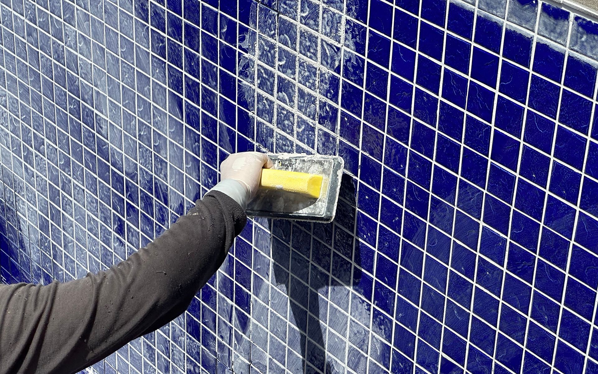 Person grouting blue tile on a wall with a yellow sponge tool, outdoors.