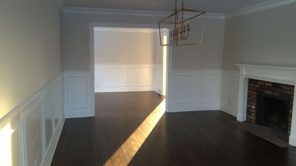 Salle à manger vide avec parquet foncé, lambris blancs et lumière du soleil inondant le sol près d'une cheminée.