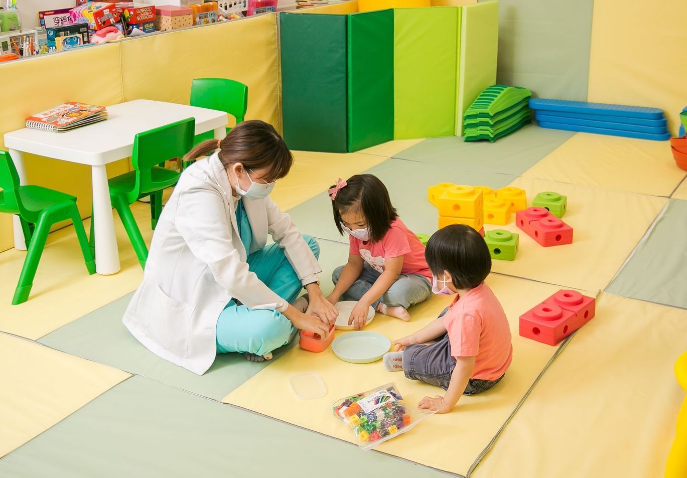 一名戴著口罩的醫護人員和兩個孩子在色彩繽紛的地板上玩耍。