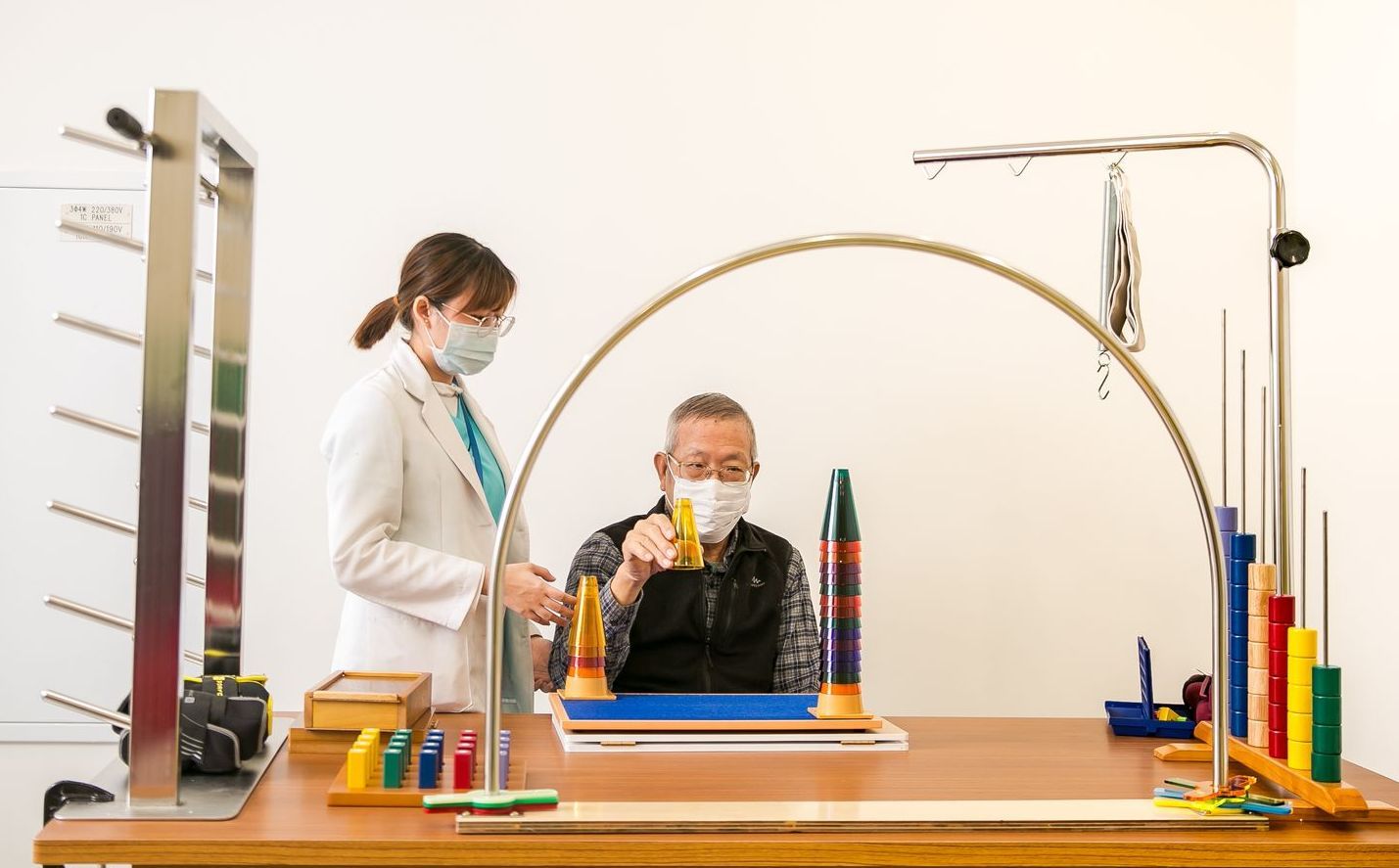 一名女子協助一名男子進行靈巧度測試，測試者使用桌上的彩色釘子和拱門。兩人都戴著口罩。