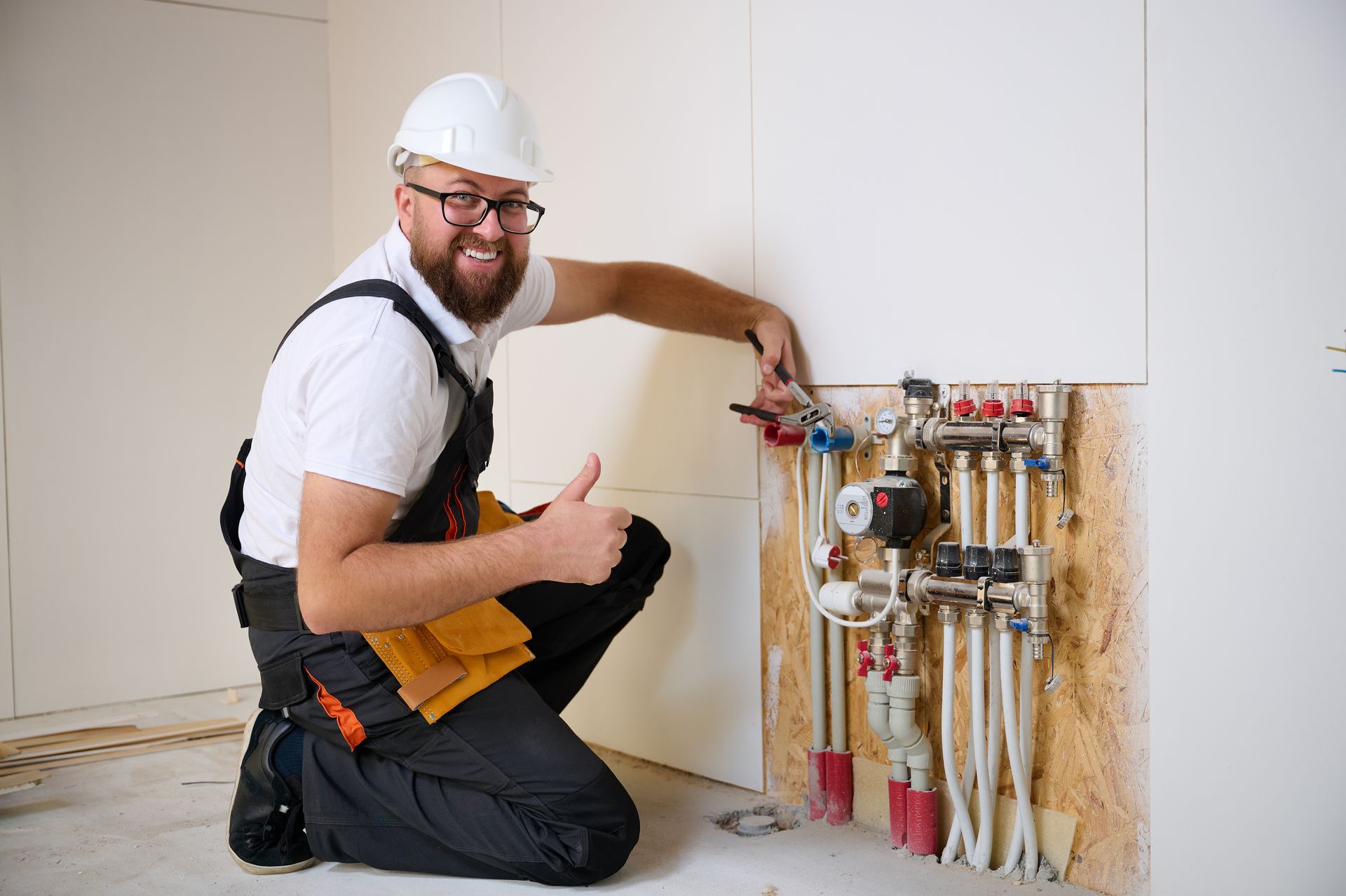 A man is kneeling down in front of a wall with pipes and giving a thumbs up.