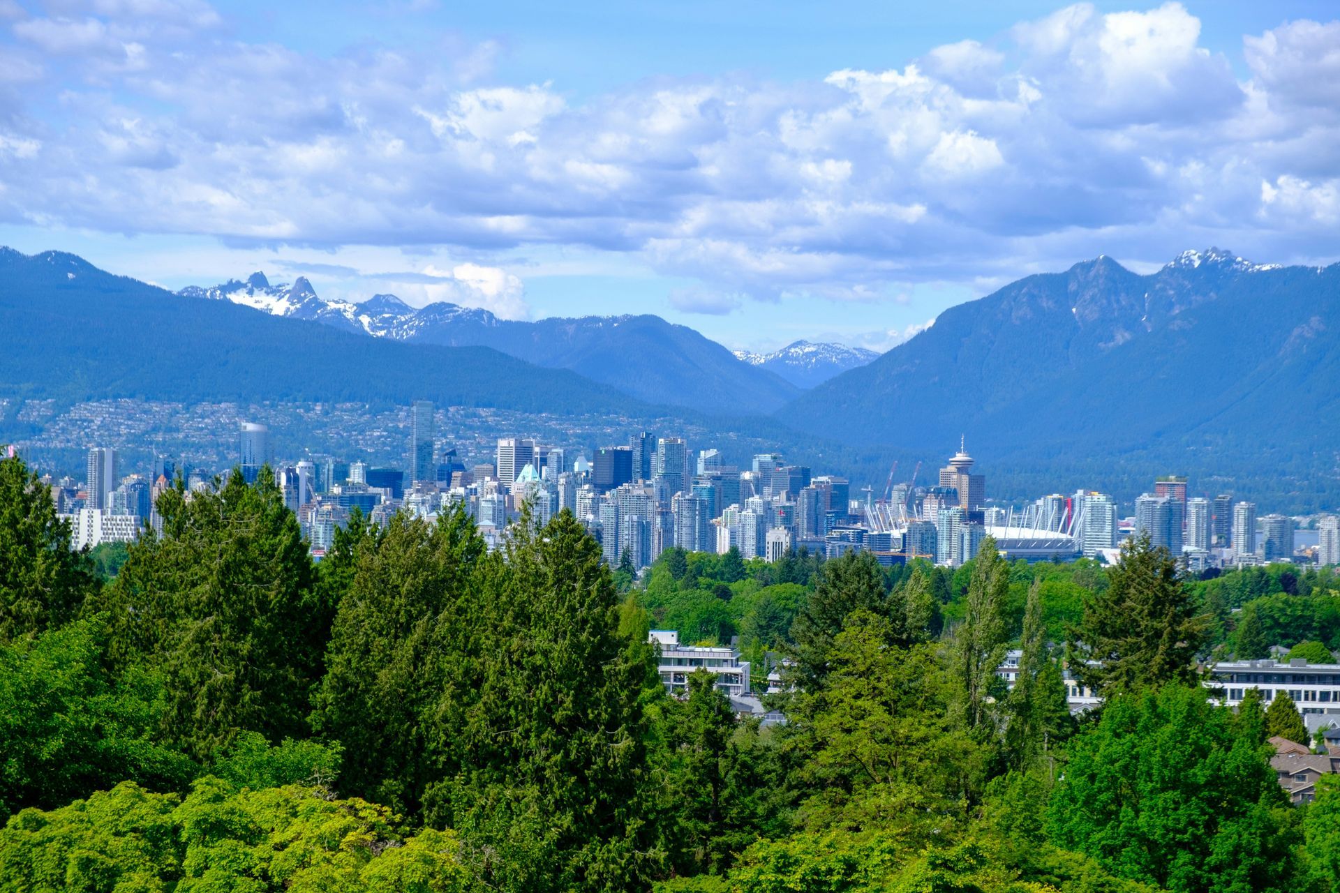 A cityscape skyline rises against a backdrop of towering, snow-capped mountains under a blue, cloudy sky.