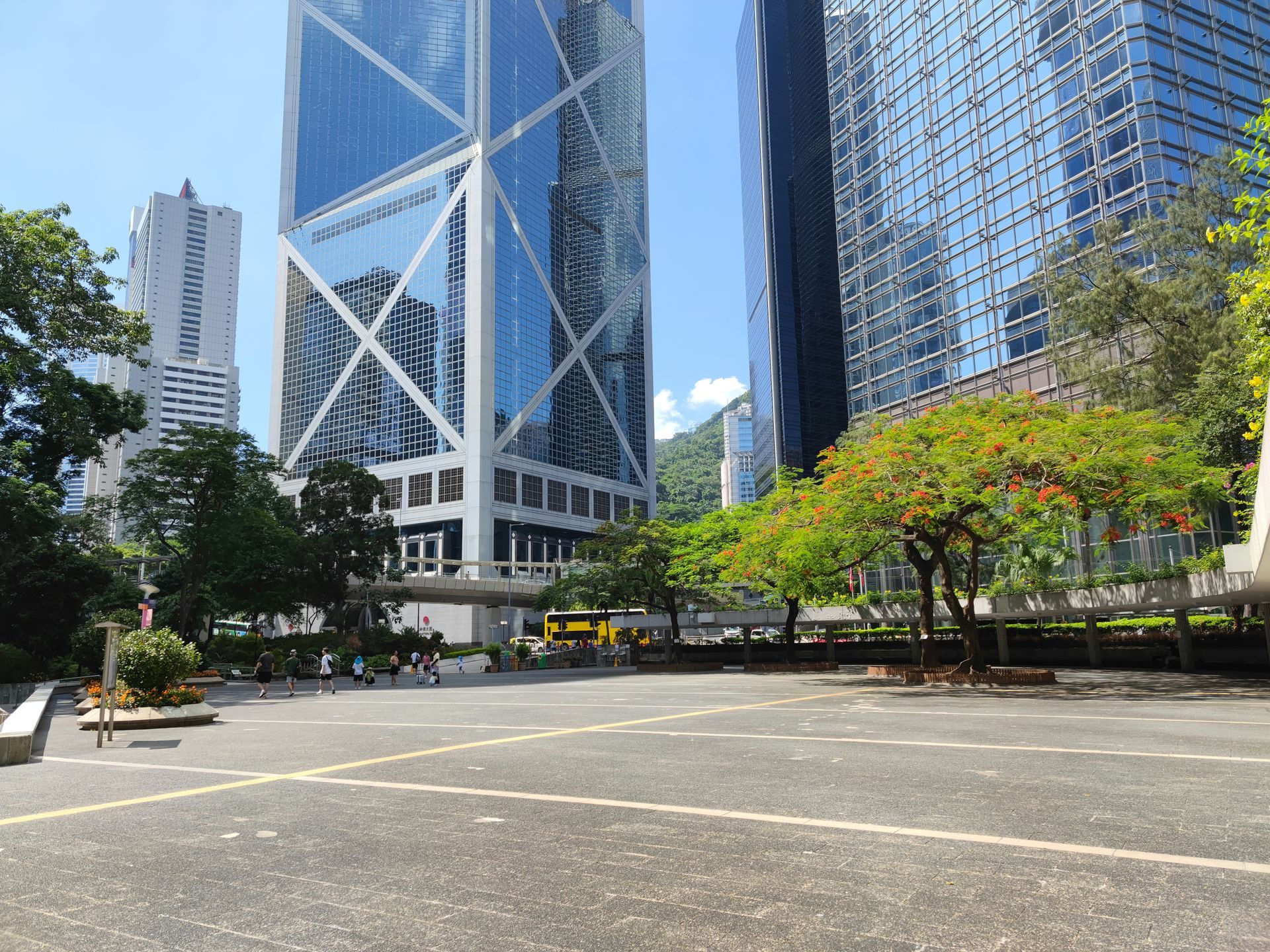 A paved plaza in Hong Kong featuring the iconic, angular Bank of China Tower beside modern glass skyscrapers and trees.