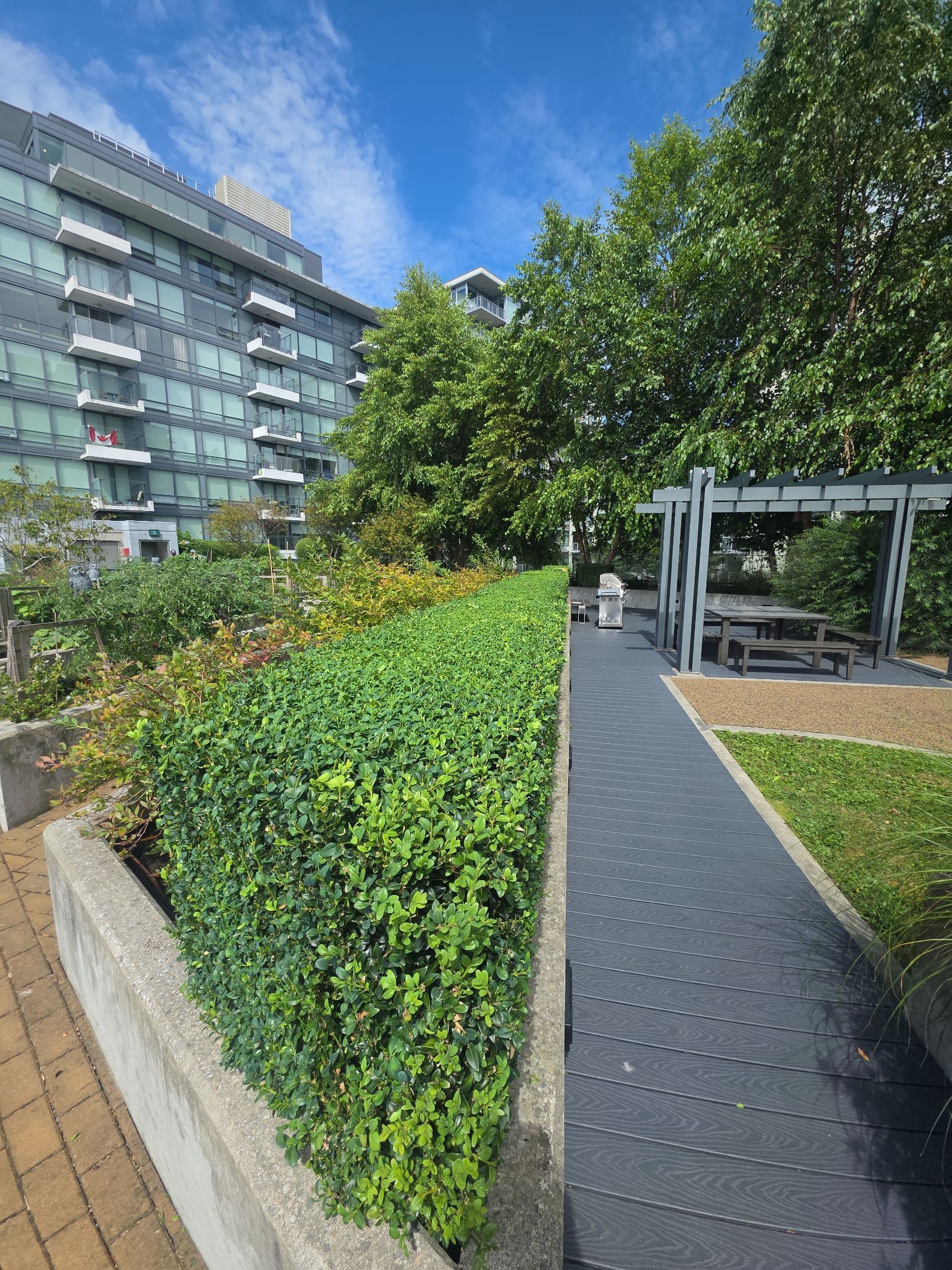 A modern apartment building overlooks a landscaped outdoor terrace with a stone planter, hedge, and a shaded seating area.