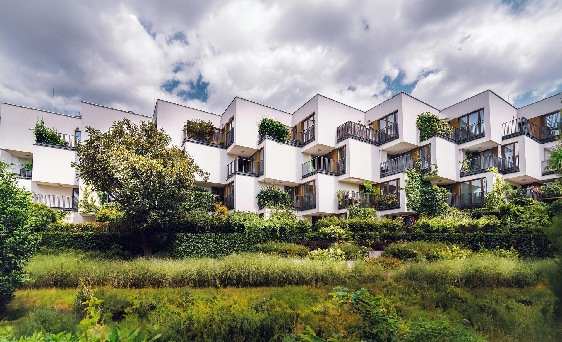 A terraced, white modern apartment complex with balconies filled with greenery, set against a cloudy blue sky.