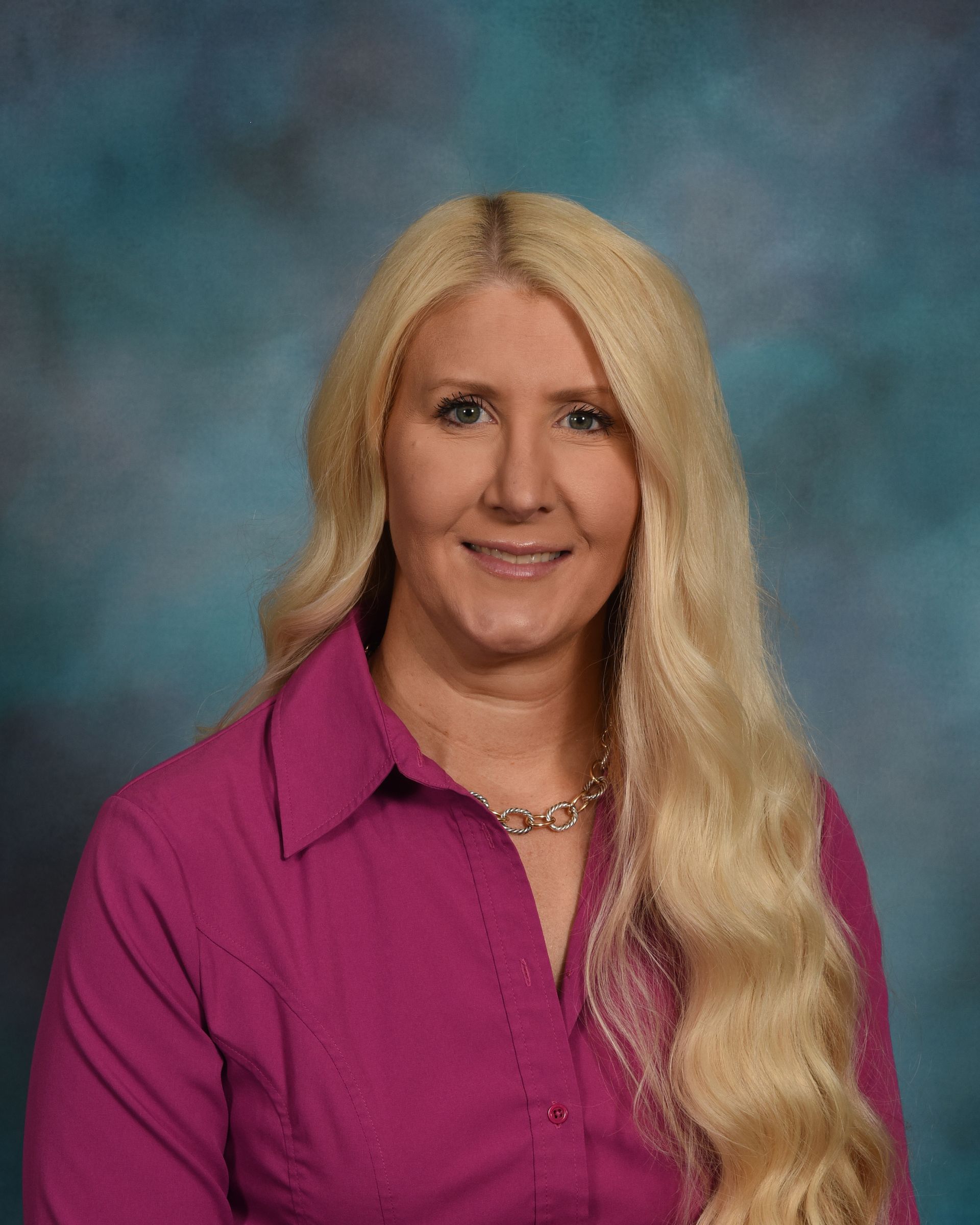 A woman with long blonde hair is wearing a pink shirt and a necklace.