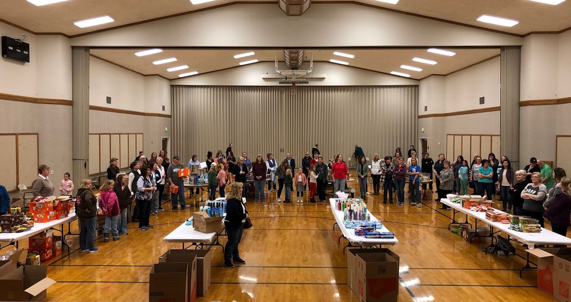 A large group of people are standing in a large room filled with tables that have snacks and supplies for snack packs.