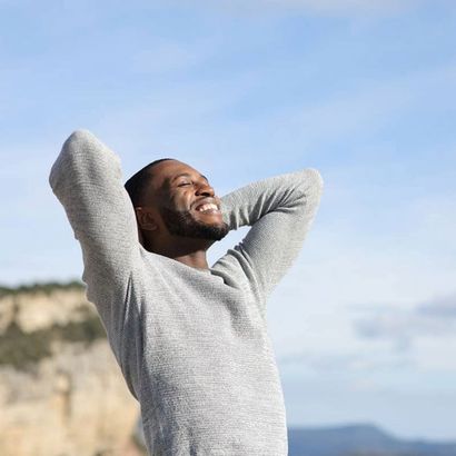 A man in a grey sweater is standing with his arms outstretched and smiling.