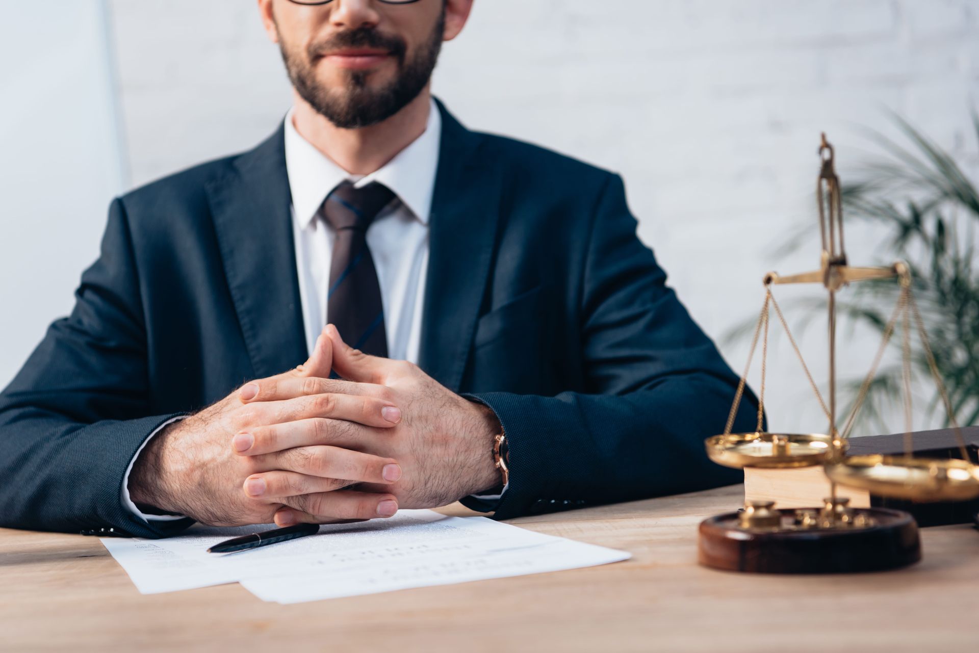 A person in a suit and tie sits at a desk with clasped hands next to a gold scale of justice.