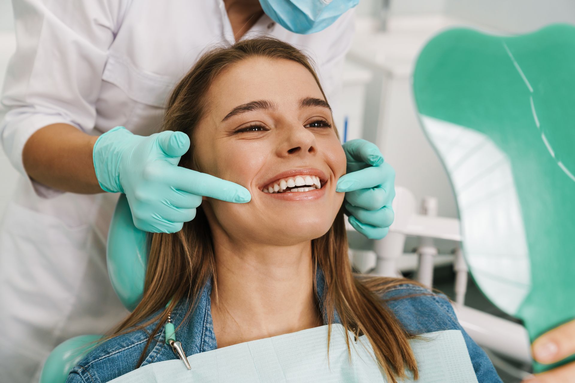 Woman at dentist smiles as professional points at teeth, holding mirror.