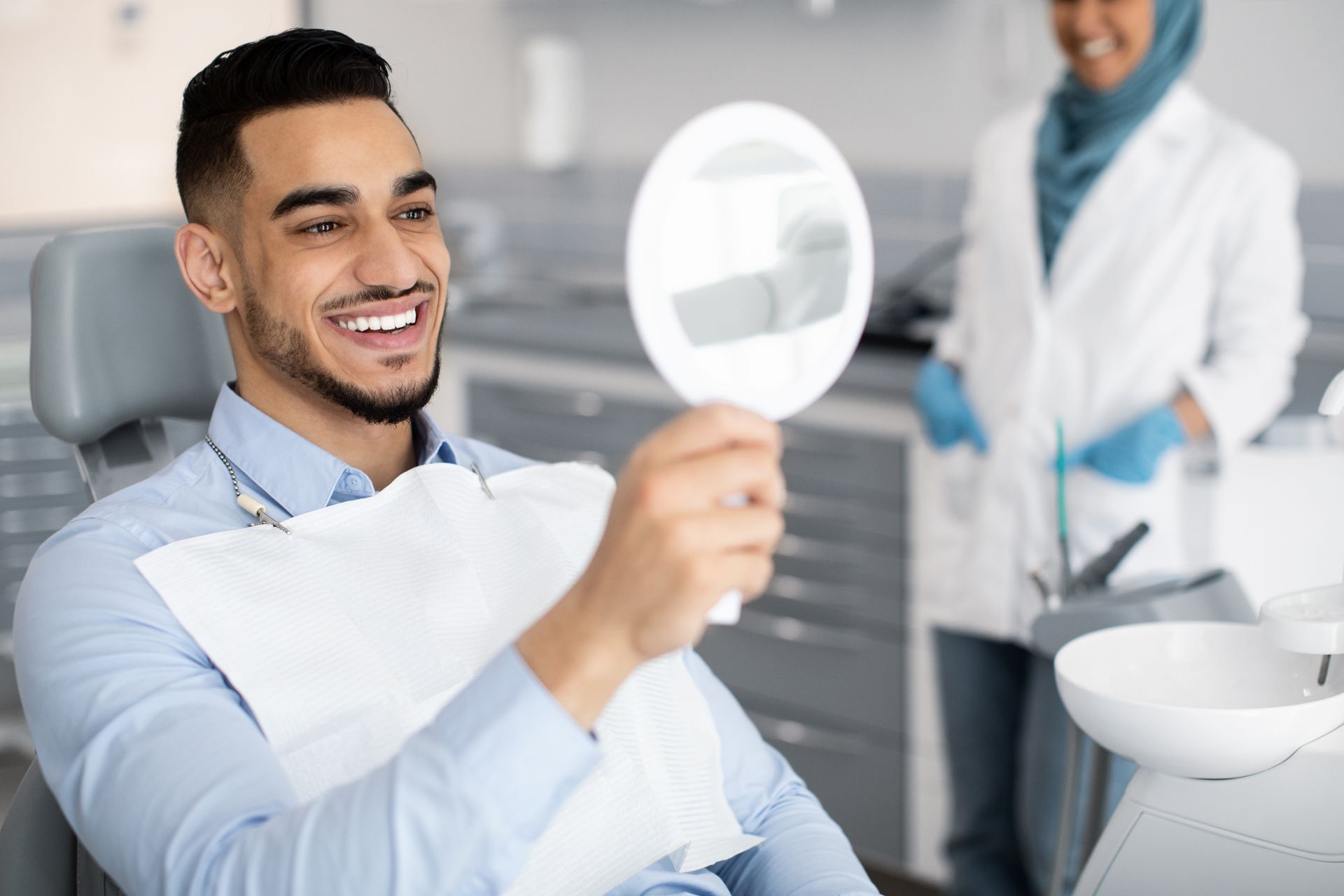 Man smiling at his teeth in a dental mirror; dentist in a hijab and lab coat in the background.