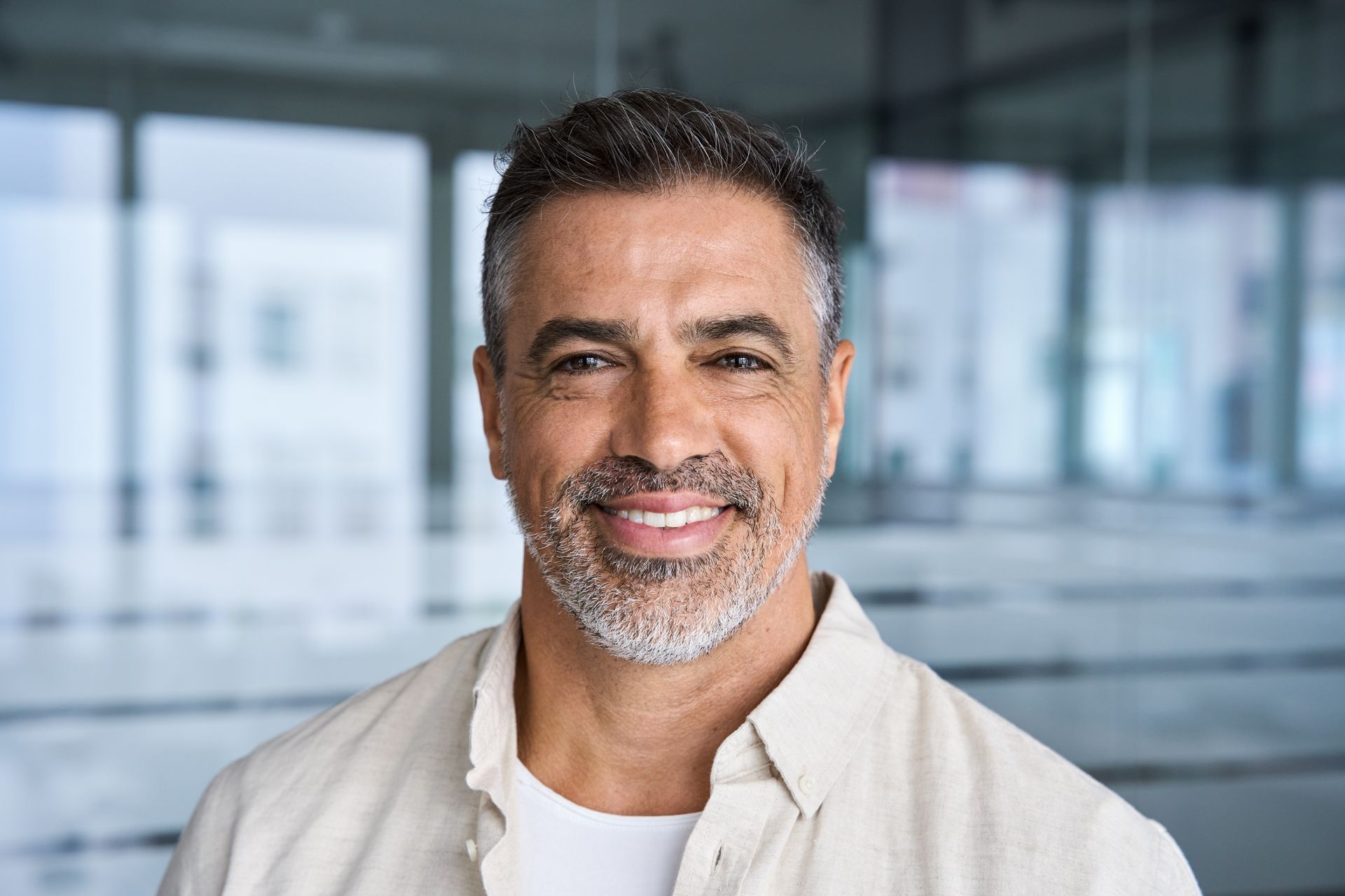 Middle-aged man with graying hair and beard smiles at camera, inside a modern building.