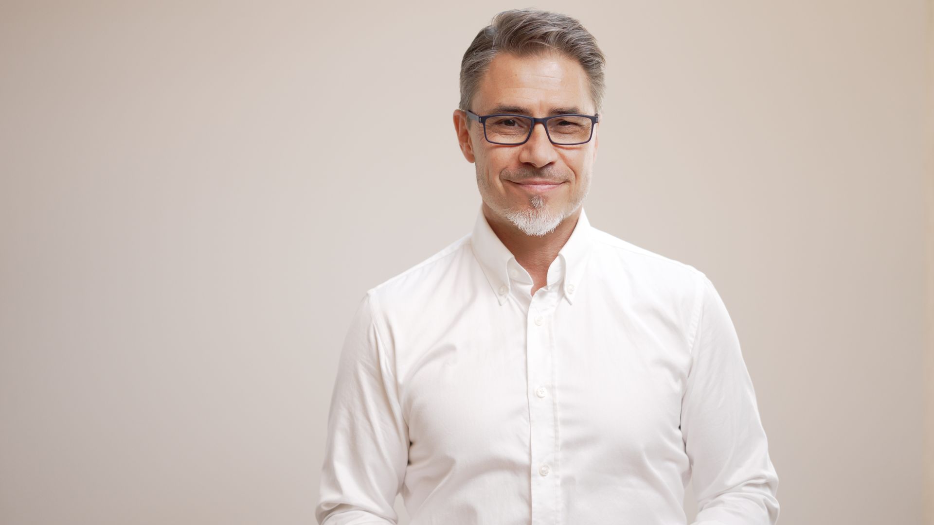 Man with graying hair and glasses in a white shirt smiles at the camera against a plain background.