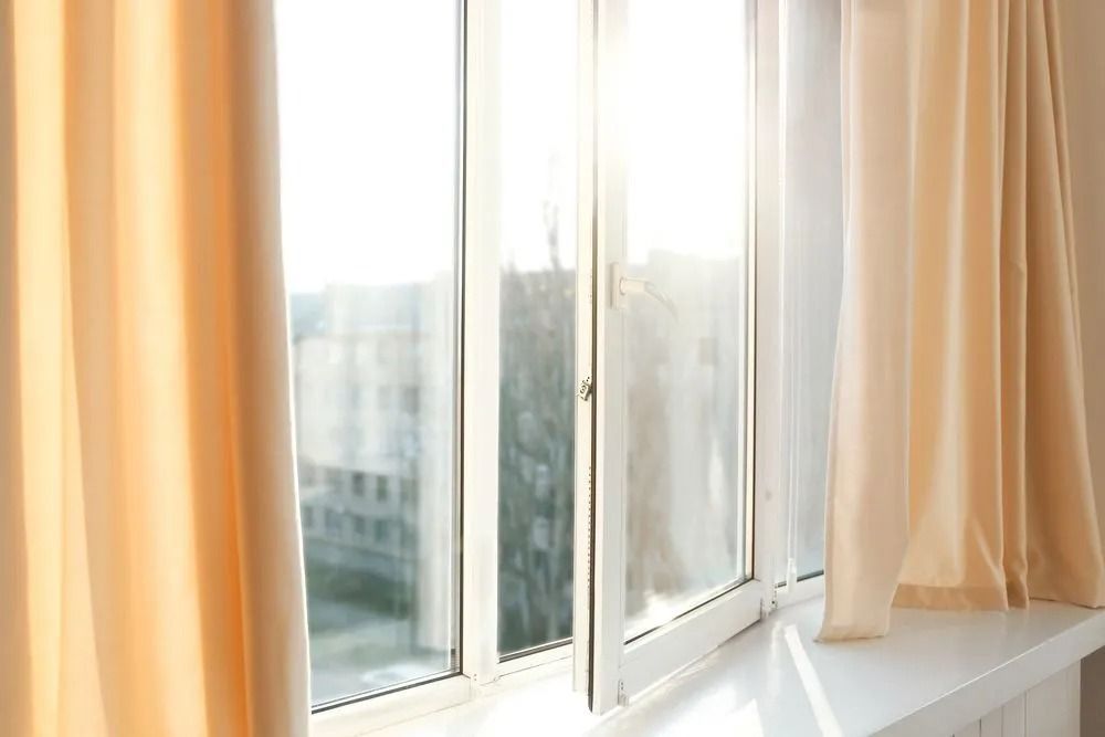 A sunlit window with beige curtains open, revealing a building and trees outdoors.