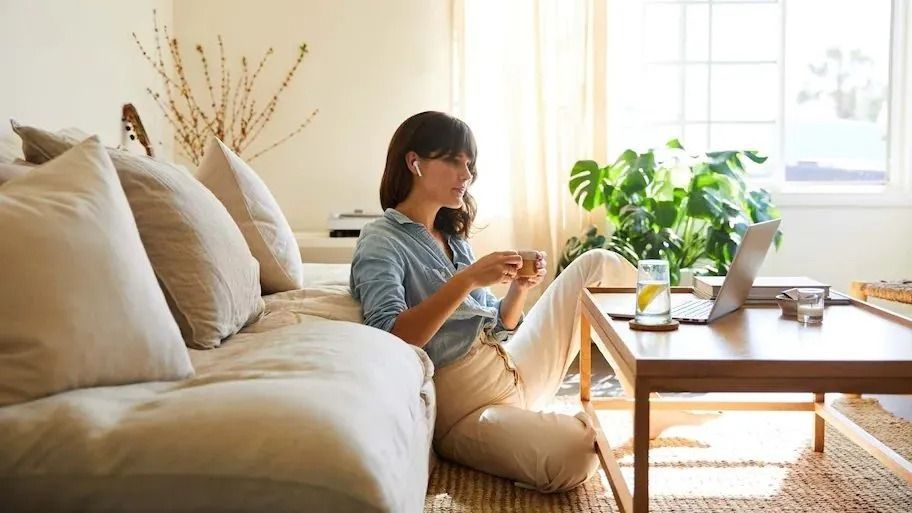 Woman working on a laptop, seated on floor in a bright living room, with a drink and a potted plant nearby.