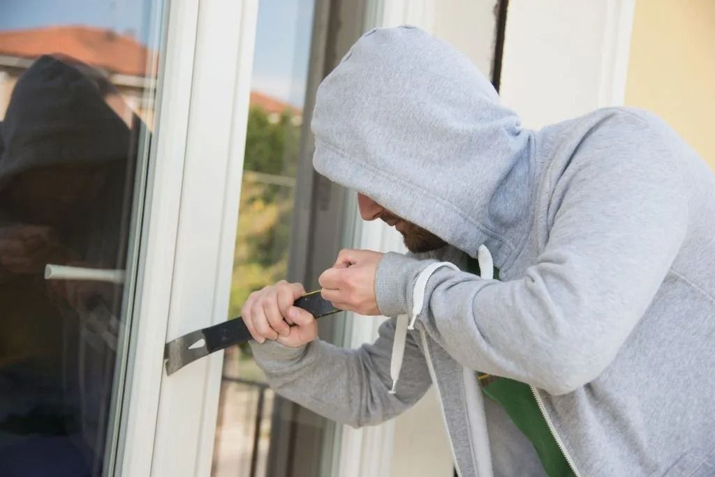 Person in a gray hoodie using a crowbar to break into a window; daylight setting.