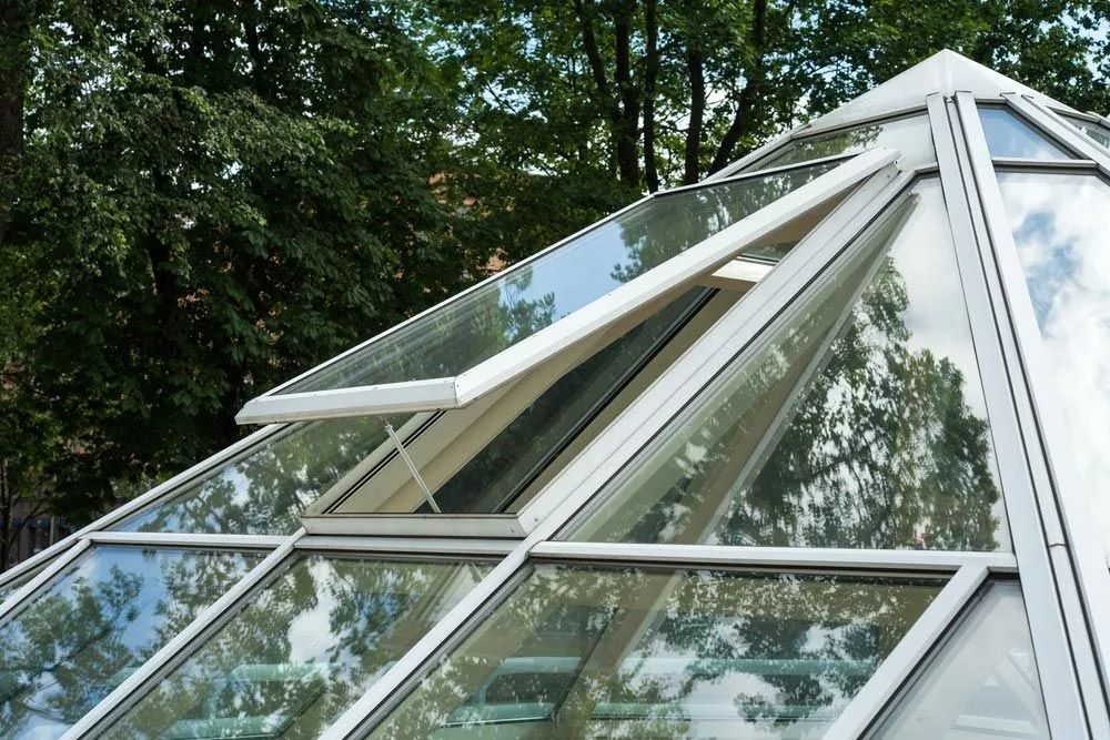 A glass pyramid with a hinged window open, revealing a view of trees; white frames and clear glass.
