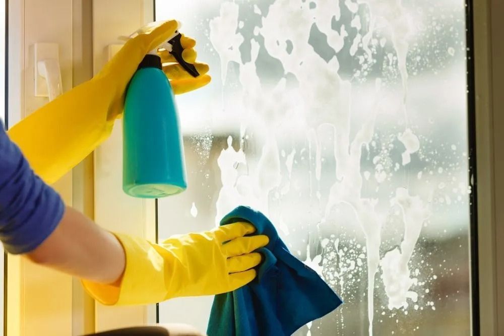 Person wearing yellow gloves sprays window cleaner and wipes a window with a blue cloth.