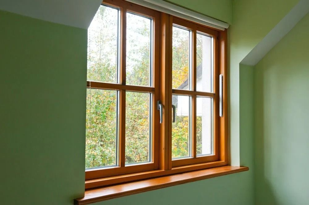 Wooden-framed window with a view of trees, set in a light-green room with a windowsill and white trim.