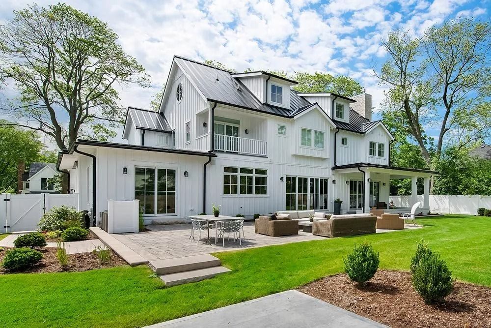 White farmhouse with a black roof and trim, large windows, a patio with seating, and a lush green lawn.