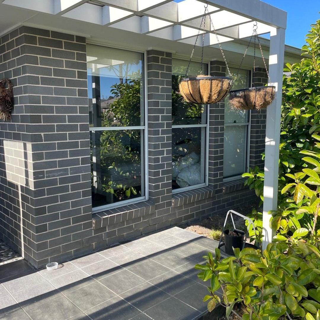 Brick building facade with large windows, a white pergola, and hanging baskets. Green foliage is visible to the right.