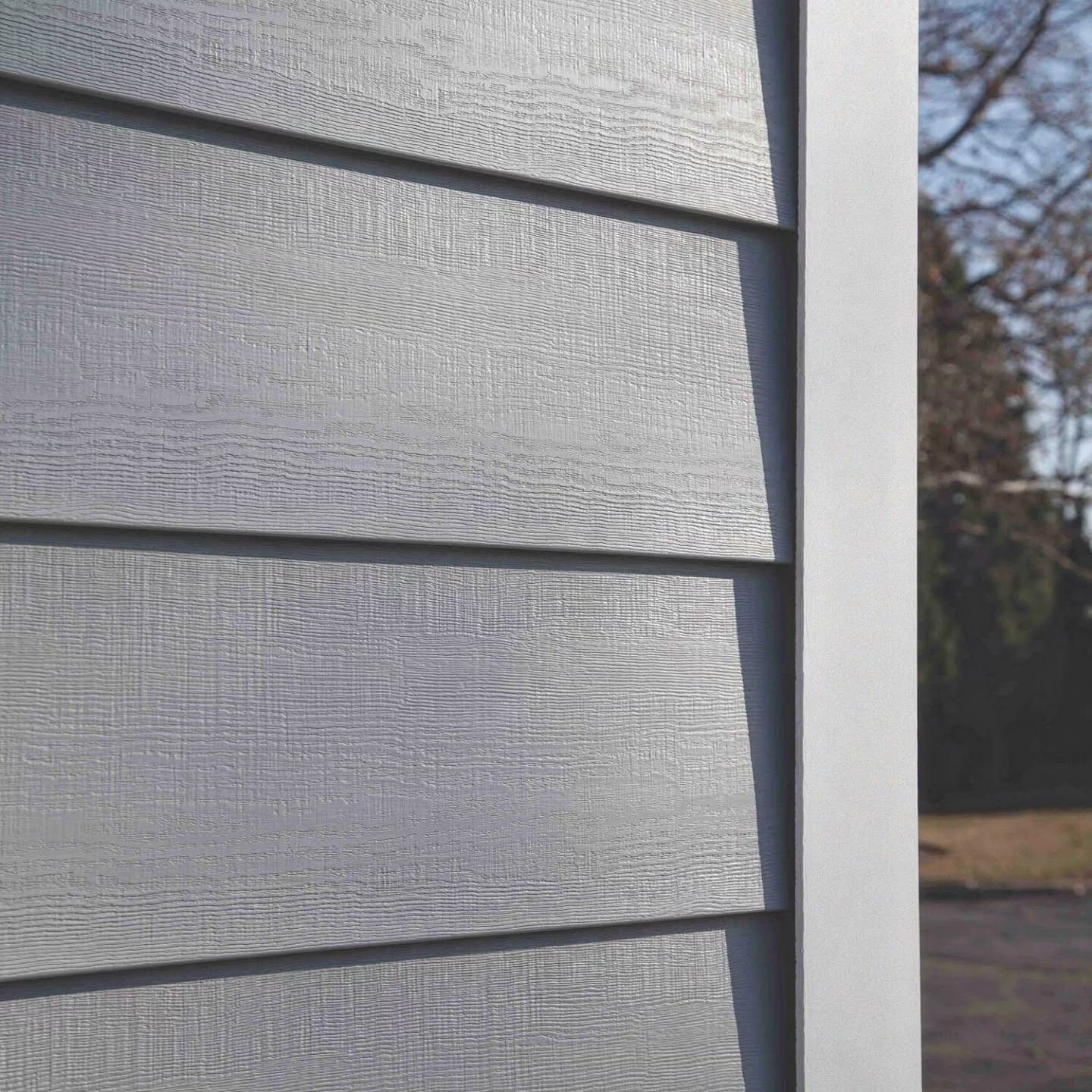 Close-up of gray siding on a building exterior with a white trim border. Sunlight creates shadows on the textured surface.