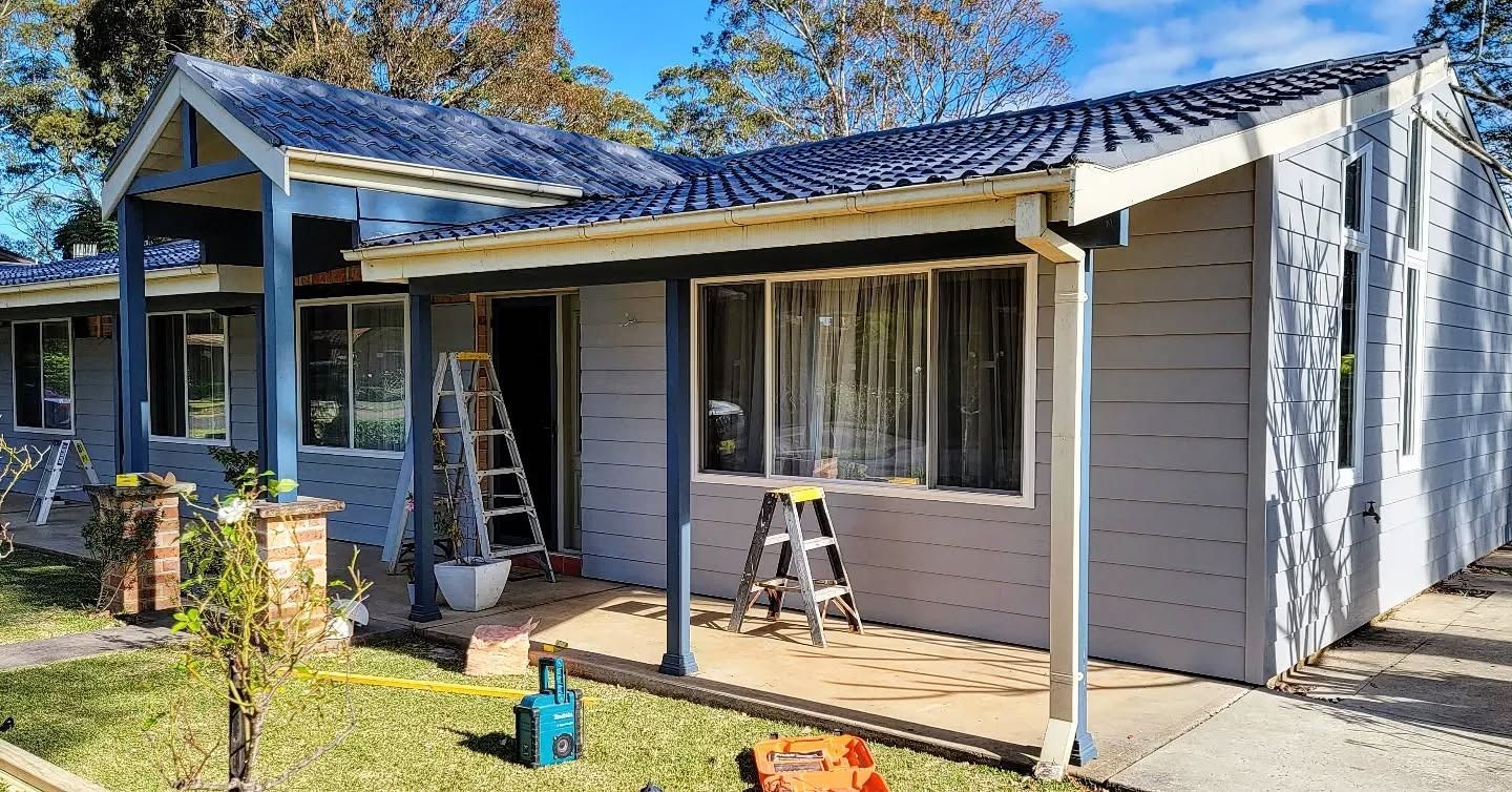 A gray house with a blue roof and porch is undergoing renovation. A ladder and tools are visible in the yard.