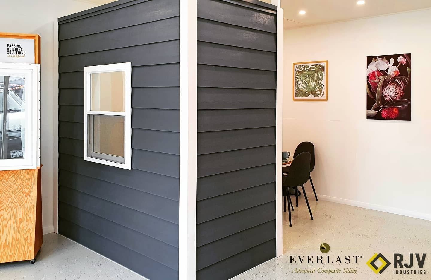 A display shows dark gray siding with a white window and trim, alongside a bright, minimalist interior with art.