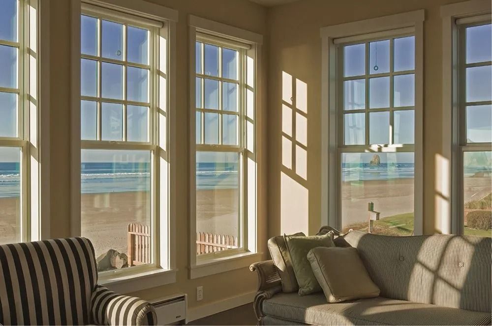 Sunlit room with tall windows offering a beach view. A striped chair and a couch with pillows are visible.