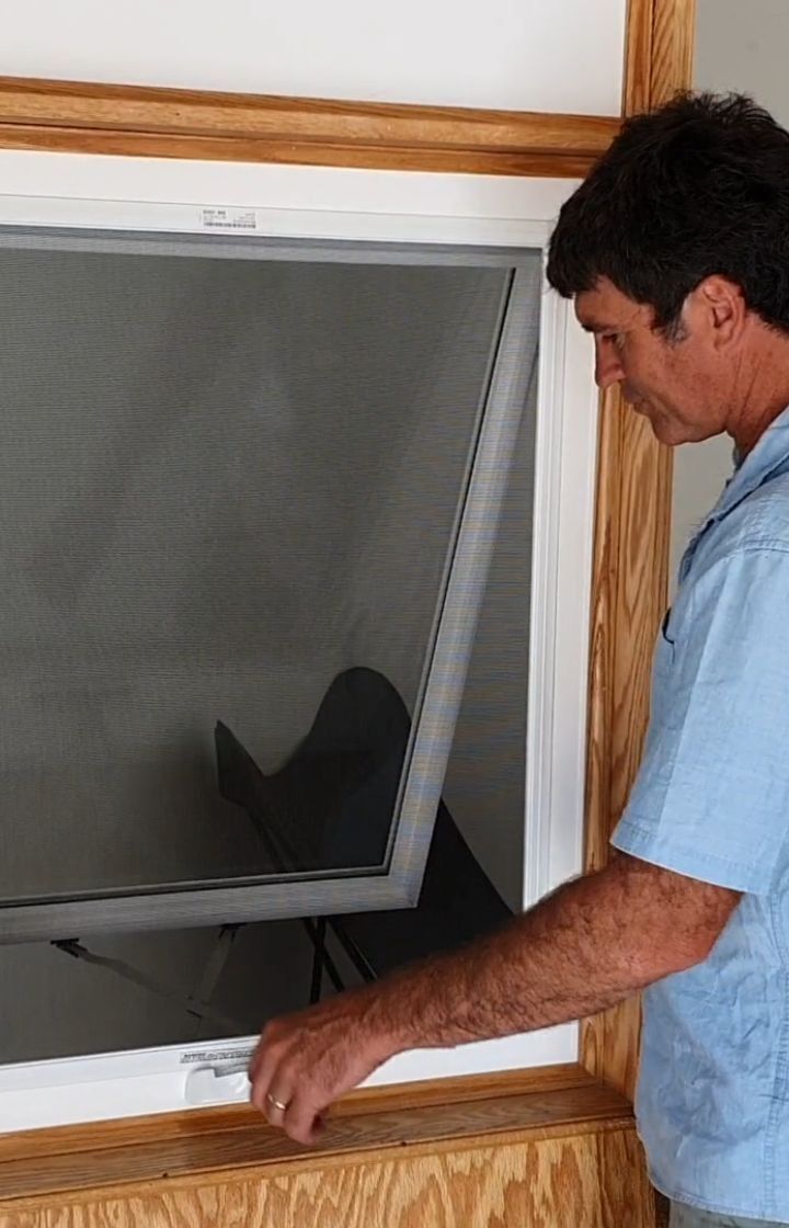 Man installing a window screen in a wooden window frame, inside a room. He is wearing a blue shirt.