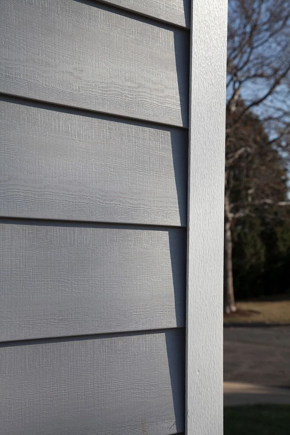 Gray siding on a building corner, showing horizontal planks with wood grain texture next to a vertical trim piece.