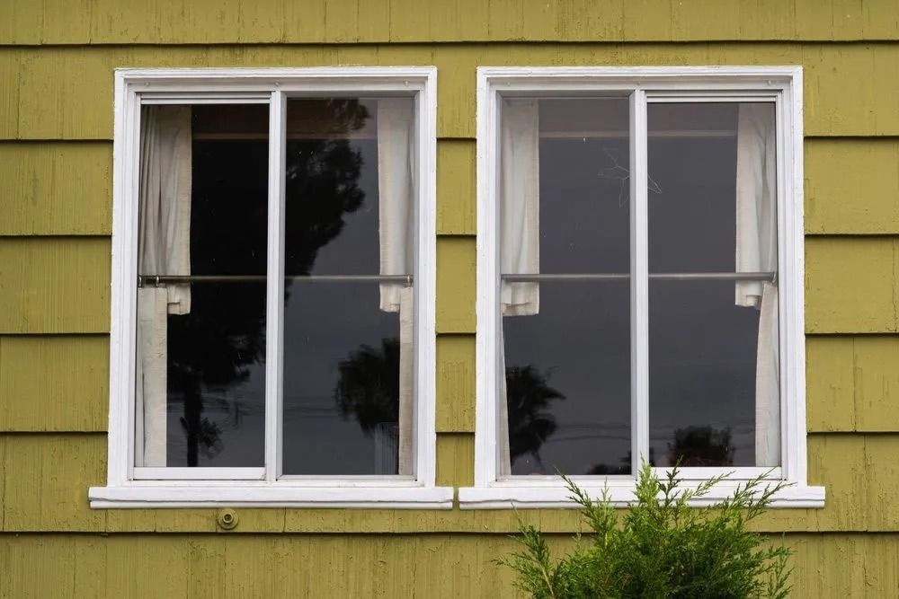 Two white-framed sliding windows on a yellow house. Sheer curtains partially cover the window panes.