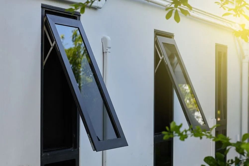 Two open, black-framed awning windows on a white building. Sunlight illuminates leaves in the background.