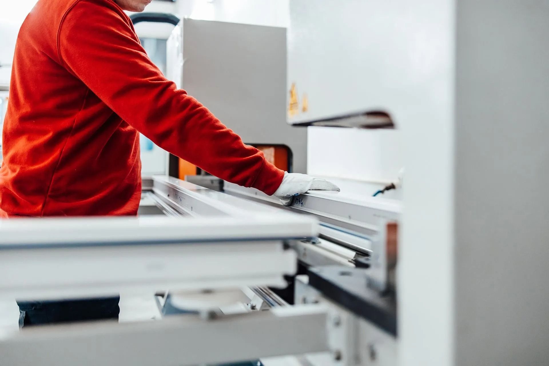 Person in red shirt and gloves works on a window frame at a manufacturing plant, using a machine.